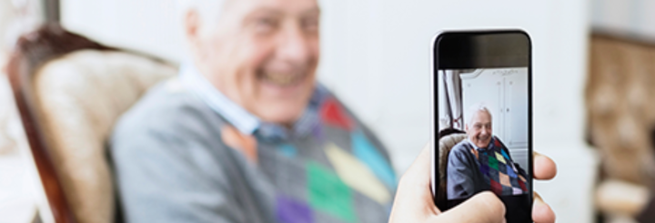 A hand holding a mobile phone, taking a picture of a smiling elderly person.